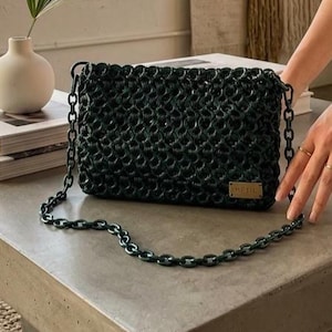 May include: Dark green handbag with a chain strap. The bag has a woven texture and a small rectangular tag with the text "ME FIL". The chain strap and the bag are the same color. The bag is sitting on a gray surface.