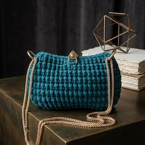 May include: Teal blue crochet handbag with a gold chain strap and clasp. The rectangular bag has a textured, woven appearance. The gold chain is draped across a dark surface. A geometric gold sculpture and a stack of books are in the background.