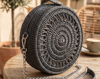 May include: A round, black woven handbag with a spiral design on the front. The bag has a silver chain strap and a small handle at the top. The bag is sitting on a wooden table.