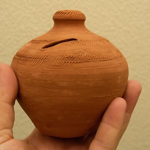 Puede incluir: Una pequeña hucha de color terracota con una ranura para monedas. El banco tiene una forma redondeada con una superficie texturizada y líneas grabadas decorativas. El banco se sostiene en una mano.