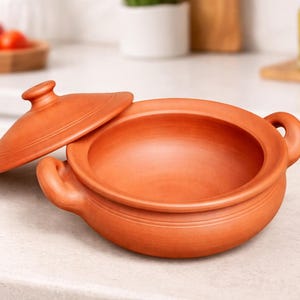 Puede incluir: Una olla de cocina de arcilla de terracota con tapa, con dos asas y forma redonda. La tapa está parcialmente abierta, revelando el interior de la olla. La olla es de color naranja cálido y terroso.