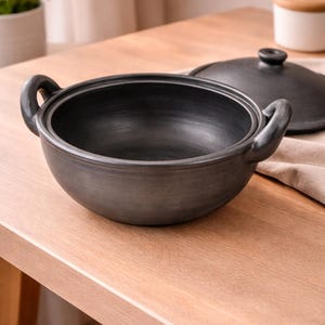 May include: A round, matte black ceramic cooking pot with two side handles and a matching circular lid. The pot features a smooth, dark finish and a wide, open bowl shape, designed for stovetop or oven use with a sturdy, heat-resistant construction.
