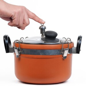 May include: An orange pressure cooker with a black handle and lid. The lid has a black knob and a silver pressure release valve. A hand is pressing down on the valve.