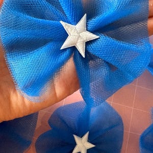 May include: Close-up of several blue tulle bows, each with a white star in the centre. The bows are arranged on a pink surface. These bows are likely for crafts or decorations.