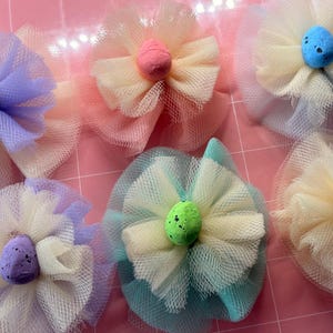 May include: Six handmade fabric flowers with tulle petals in pastel shades, each with a speckled egg-shaped centre. The flowers are arranged on a pink surface, ideal for Easter or spring crafts.