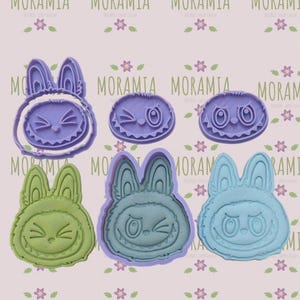 May include: A collection of colorful cookie cutters in the shape of cartoon bunnies and winking faces. The cutters are in shades of purple, green, and blue. The brand name "MORAMIA" is repeated across the top and bottom of the image.