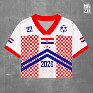 May include: A white short-sleeve jersey with a red V-neck collar and red sleeve cuffs. The front features a red and white checkered pattern, a blue chevron design, the number 22, a soccer ball icon, the Croatian flag, and the text 2026.