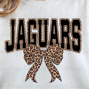 May include: White sweatshirt with the word "JAGUARS" in black block letters with a leopard print fill. A leopard print bow is centered below the word. The sweatshirt is a casual style.