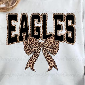 May include: White sweatshirt with the word "EAGLES" in black block letters with a leopard print fill. A leopard print bow is centered below the word. The sweatshirt is a casual style.