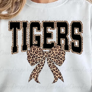 May include: White sweatshirt with the word "TIGERS" in black block letters with a leopard print fill. A leopard print bow is centered below the word. The sweatshirt is a casual style.