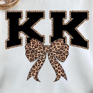 以下が含まれることがあります： 黒い「KK」の文字とヒョウ柄の縁取りが特徴の白いスウェットシャツ。ヒョウ柄のリボンが文字の下に配置されています。スウェットシャツはカジュアルなスタイルです。