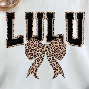 Könnte beinhalten: Weißes Sweatshirt mit dem Namen "LULU" in großen, schwarzen Blockbuchstaben mit Leopardenmuster-Umrandung. Eine große Schleife mit Leopardenmuster befindet sich mittig unter den Buchstaben. Das Sweatshirt hat einen lässigen Stil.