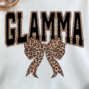 May include: White sweatshirt with the word "GLAMMA" in large, black, leopard print letters. A leopard print bow is centered below the word. The sweatshirt is a casual style.