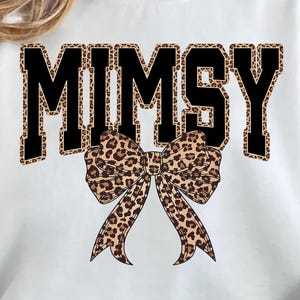 Op de afbeelding: Witte sweatshirt met het woord "MIMSY" in zwarte blokletters met een luipaardprint. Een grote luipaardprint strik is gecentreerd onder de tekst. De sweatshirt heeft een casual stijl.