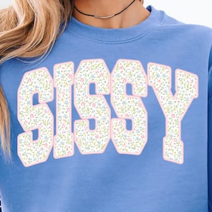 May include: A light blue sweatshirt with the word "SISSY" in large, floral-patterned letters. The letters have a white background with a pink outline, and the sweatshirt has long sleeves.