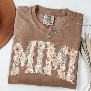 May include: A brown Comfort Colors t-shirt with the word "MIMI" in large, floral-filled letters. A silver necklace with a small pendant rests on the shirt. The shirt is folded, with a brown hat visible in the background.