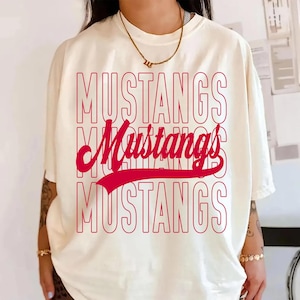 May include: Cream-colored t-shirt with the word "MUSTANGS" in red, stacked three times, with a cursive "Mustangs" in the middle. The shirt has short sleeves and a relaxed fit.