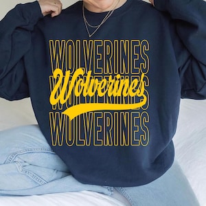 Op de afbeelding: Marineblauw sweatshirt met het woord "Wolverines" in een gele, gestapelde opmaak. Het woord "Wolverines" wordt driemaal herhaald, met een gele swoosh onder het middelste woord. Een casual kledingstuk.