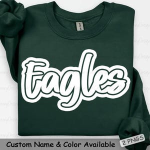 May include: A dark green sweatshirt with the word "Eagles" in a white, stylized font. The text is outlined in black. The sweatshirt has a crew neck and long sleeves. The text "Custom Name & Color Available" is at the bottom.