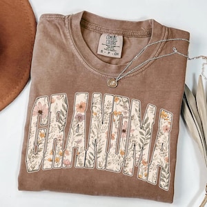 Op de afbeelding: Een bruin Comfort Colors t-shirt met het woord "GRANDMA" in een bloemenontwerp. Een zilveren ketting met een hanger is over het shirt gedrapeerd. Het shirt is een neutrale kleur en heeft een vintage look.