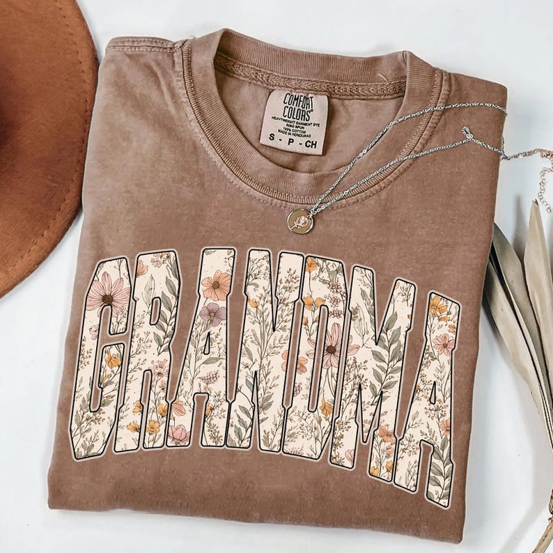 Op de afbeelding: Een bruin Comfort Colors t-shirt met het woord "GRANDMA" in een bloemenontwerp. Een zilveren ketting met een hanger is over het shirt gedrapeerd. Het shirt is een neutrale kleur en heeft een vintage look.