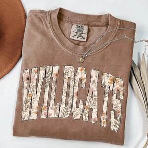 Puede incluir: Una camiseta marr&oacute;n con la palabra "WILDCATS" en letras grandes llenas de flores. La camiseta tiene cuello redondo y mangas cortas. Un collar plateado con un peque&ntilde;o colgante est&aacute; drapeado sobre la camiseta. La camiseta es talla S.