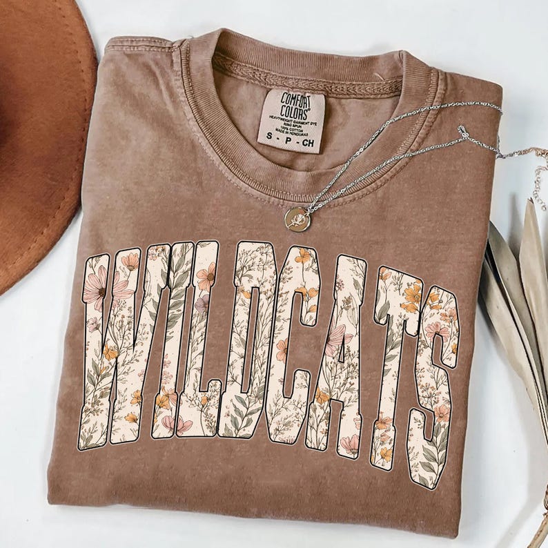 Puede incluir: Una camiseta marr&oacute;n con la palabra "WILDCATS" en letras grandes llenas de flores. La camiseta tiene cuello redondo y mangas cortas. Un collar plateado con un peque&ntilde;o colgante est&aacute; drapeado sobre la camiseta. La camiseta es talla S.