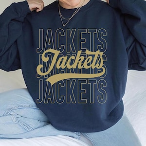 May include: Navy blue sweatshirt with the word "JACKETS" in a gold, layered font. The word is repeated three times, with a stylized script font in the middle. The sweatshirt is a casual style.