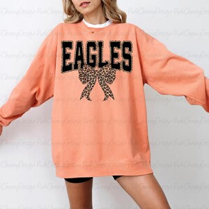May include: A coral-coloured sweatshirt featuring the word "EAGLES" in black block letters with a leopard print trim. Below the word is a leopard print bow. The sweatshirt has long sleeves.