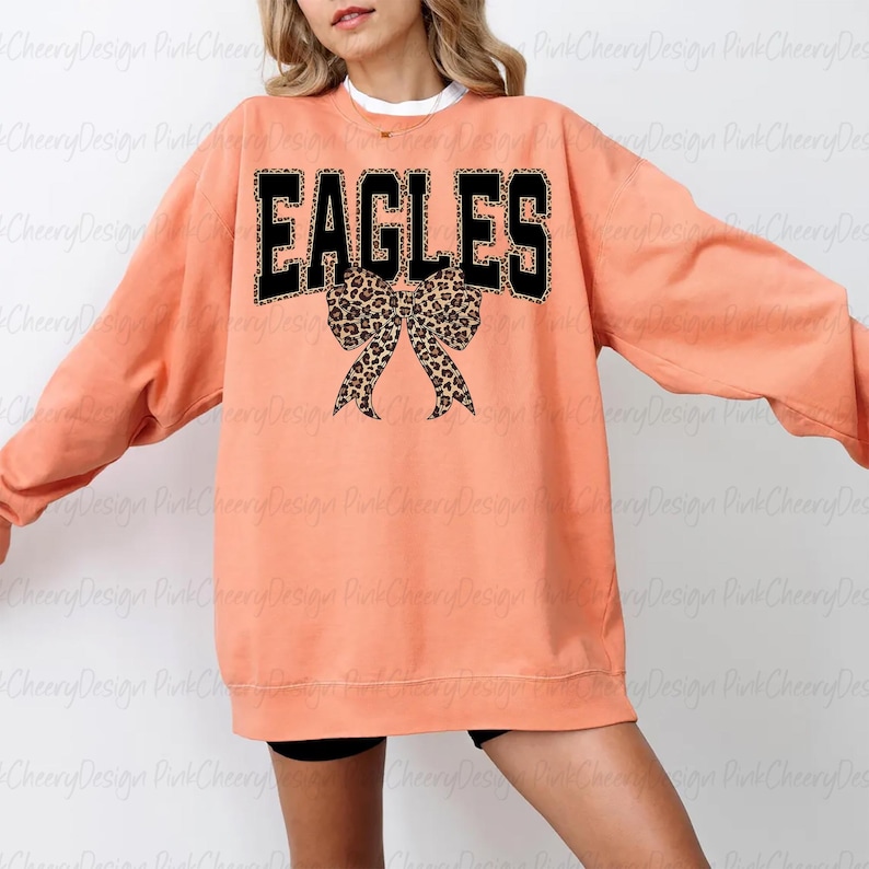 May include: A coral-coloured sweatshirt featuring the word "EAGLES" in black block letters with a leopard print trim. Below the word is a leopard print bow. The sweatshirt has long sleeves.