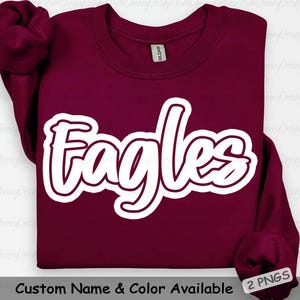 May include: A maroon sweatshirt with the word "Eagles" in a white, outlined script font. The sweatshirt has long sleeves and a crew neck. The text "Custom Name & Color Available" is at the bottom.