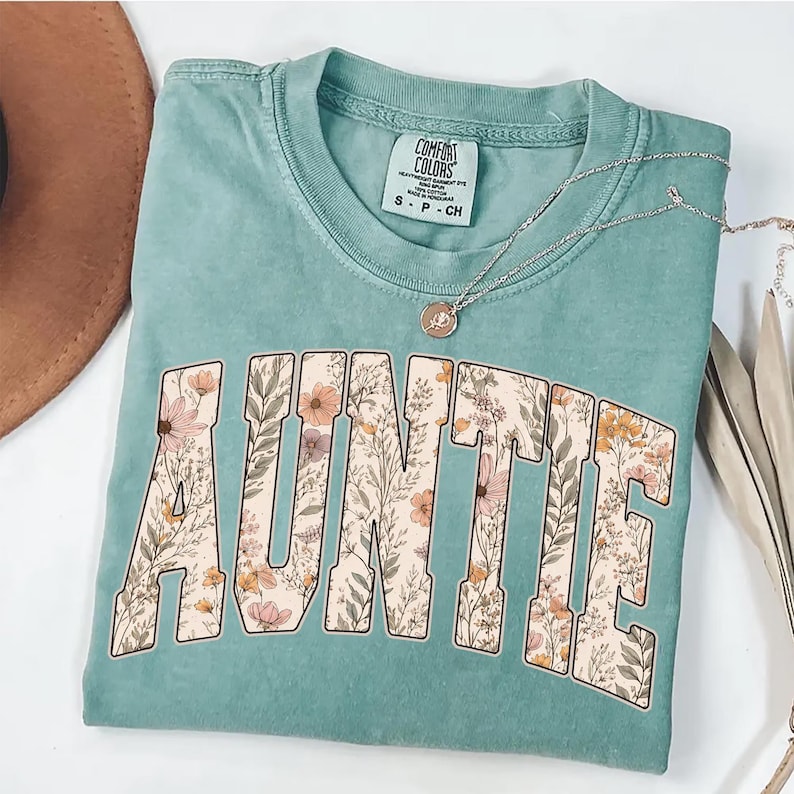 May include: A seafoam green t-shirt with the word "AUNTIE" in large, floral-filled letters. The shirt has a crew neck and short sleeves. A silver necklace with a pendant is draped across the shirt.