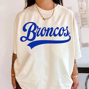 Puede incluir: Camiseta color crema con la palabra "Broncos" en una fuente azul negrita en la parte delantera. La palabra está arqueada y tiene una línea azul debajo. La camiseta tiene mangas cortas y un corte holgado.