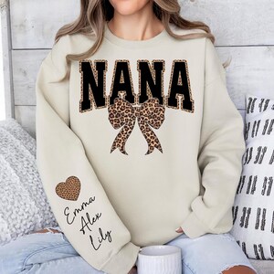 Puede incluir: Sudadera color crema con la palabra "NANA" en letras negras grandes con borde de estampado de leopardo. Un lazo con estampado de leopardo est&aacute; centrado debajo de la palabra. La manga izquierda tiene un coraz&oacute;n con estampado de leopardo y los nombres "Emma, Alex, Lily".