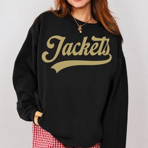 May include: Black sweatshirt with the word "Jackets" in gold script across the chest. The sweatshirt has a crew neck and long sleeves. The text has a shadow effect. The model is wearing a gold necklace and red and white checkered pants.