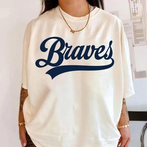 以下が含まれることがあります： 前面にネイビーブルーの筆記体で「Braves」と書かれたクリーム色のTシャツ。単語はネイビーブルーの水平バーの上にアーチ状になっています。シャツは半袖で、ゆったりとしたフィット感です。