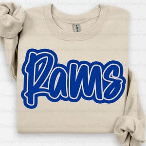 Pode incluir: Camisola bege com a palavra "Rams" em azul vibrante, contornada a branco. O design é um gráfico arrojado e apelativo. A camisola tem gola redonda e mangas compridas.