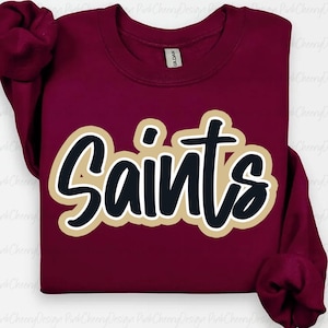 以下が含まれることがあります： 黒、金、白のデザインで「Saints」の文字が入った、バーガンディのクルーネックのトレーナー。トレーナーは長袖で、Gildanのタグが付いています。テキストは太字の筆記体で、金の縁取りがされています。