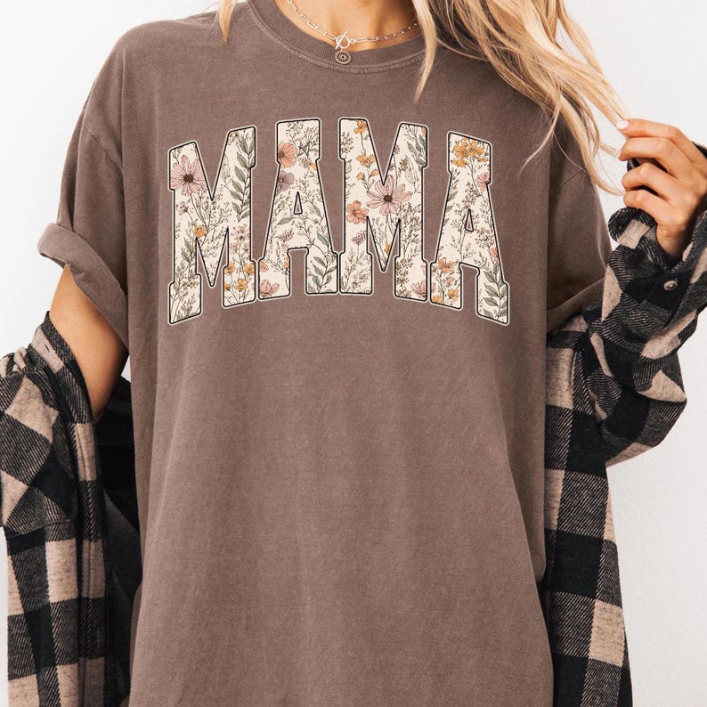 May include: A brown t-shirt with the word "MAMA" in a floral design. The letters are filled with a pattern of flowers and leaves in shades of pink, yellow, and green. The shirt has short sleeves and a crew neck.