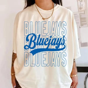 Peut inclure: T-shirt crème avec le mot "BLUEJAYS" en bleu, répété trois fois. Le "Bluejays" du milieu est en écriture cursive avec un trait bleu en dessous. Un collier doré est visible.