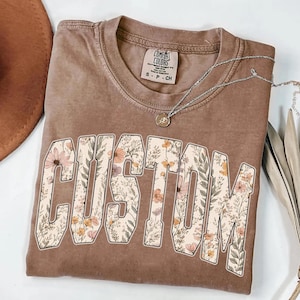 Puede incluir: Una camiseta marr&oacute;n con la palabra "CUSTOM" en letras grandes y florales. La camiseta tiene cuello redondo y mangas cortas. Un collar plateado con un peque&ntilde;o colgante est&aacute; sobre la camiseta.