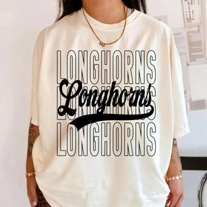Puede incluir: Camiseta color crema con la palabra "LONGHORNS" en un dise&ntilde;o apilado. La palabra "Longhorns" se repite tres veces en una fuente negra en negrita, con una versi&oacute;n de gui&oacute;n estilizada en el centro. Una l&iacute;nea negra curva subraya el gui&oacute;n.