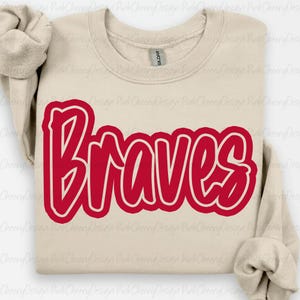 Op de afbeelding: Een beige crewneck sweatshirt met het woord "Braves" in vetgedrukte rode letters. Het woord heeft een witte omlijning. De sweatshirt heeft lange mouwen en een geribbelde halslijn.