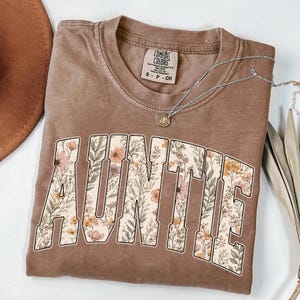 May include: A brown t-shirt with the word "AUNTIE" in large, floral-filled letters. The shirt has a crew neck and short sleeves. A silver necklace with a small pendant is draped across the shirt.