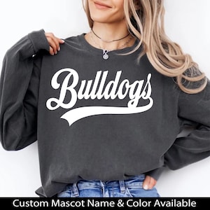 K&ouml;nnte beinhalten: Ein dunkelgraues Sweatshirt mit dem wei&szlig;en Schriftzug "Bulldogs" auf der Vorderseite. Das Sweatshirt hat lange &Auml;rmel und einen Rundhalsausschnitt. Der Text "Custom Mascot Name & Color Available" befindet sich unten.
