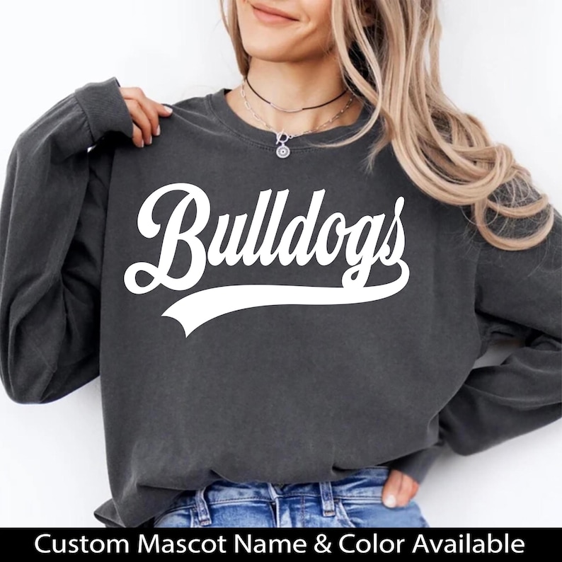K&ouml;nnte beinhalten: Ein dunkelgraues Sweatshirt mit dem wei&szlig;en Schriftzug "Bulldogs" auf der Vorderseite. Das Sweatshirt hat lange &Auml;rmel und einen Rundhalsausschnitt. Der Text "Custom Mascot Name & Color Available" befindet sich unten.