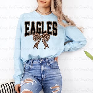 May include: A pale blue long-sleeved top featuring the word "EAGLES" in black block letters with a leopard print fill. A leopard print bow is centered below the word. The top is worn with blue jeans.
