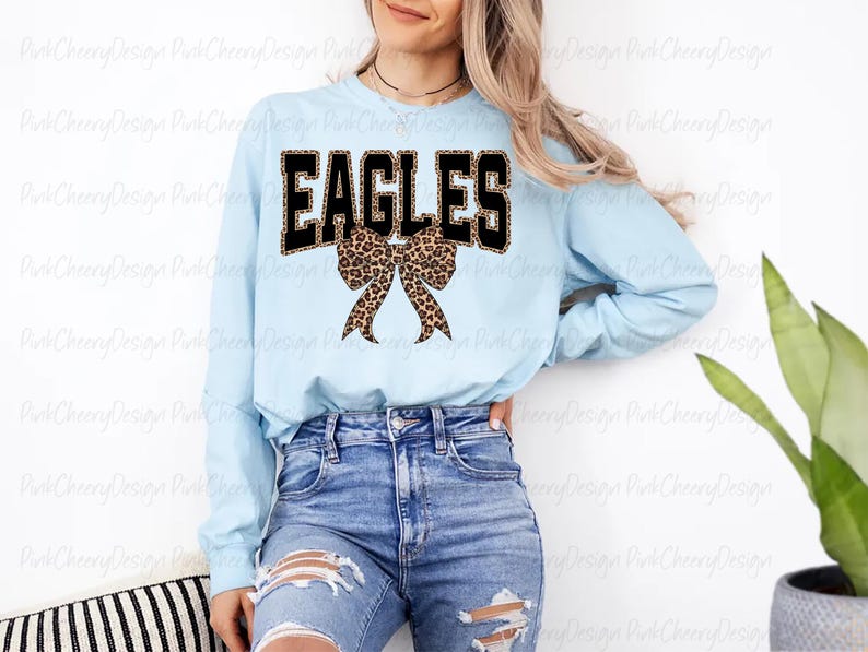May include: A pale blue long-sleeved top featuring the word "EAGLES" in black block letters with a leopard print fill. A leopard print bow is centered below the word. The top is worn with blue jeans.