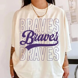 Peut inclure: T-shirt crème avec le mot "BRAVES" dans un design superposé. Les mots du haut et du bas sont soulignés en violet clair, tandis que le mot du milieu "Braves" est en police cursive violet foncé.