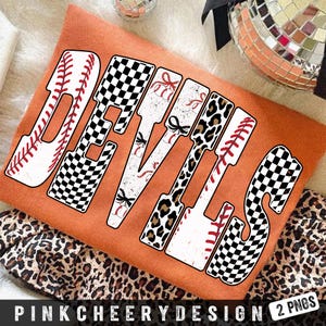 Puede incluir: Una camiseta naranja con la palabra "DEVILS" en una fuente audaz y decorativa. Las letras presentan costuras de béisbol, patrones de cuadros y estampados de leopardo. La parte inferior de la camiseta está parcialmente cubierta por una tela con estampado de leopardo.